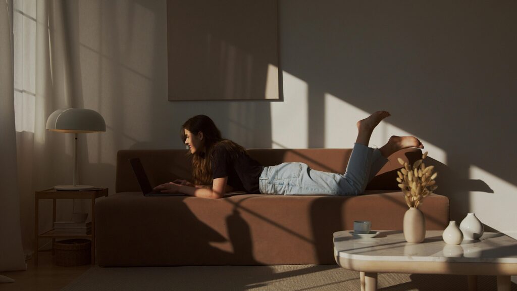 A young woman lying on a sofa using a laptop, enjoying a cozy and relaxed afternoon indoors.